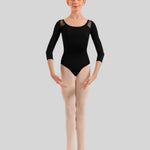A young ballerina stands facing forward wearing a black three-quarter sleeve leotard with sheer shoulder panels and light beige tights against a solid gray background. 