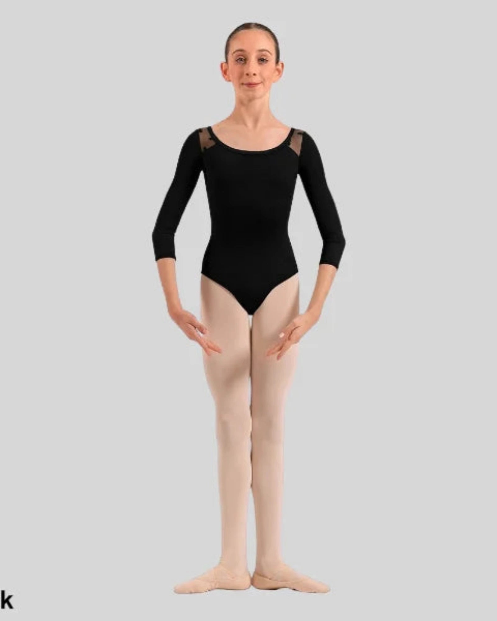 A young ballerina stands facing forward wearing a black three-quarter sleeve leotard with sheer shoulder panels and light beige tights against a solid gray background. 