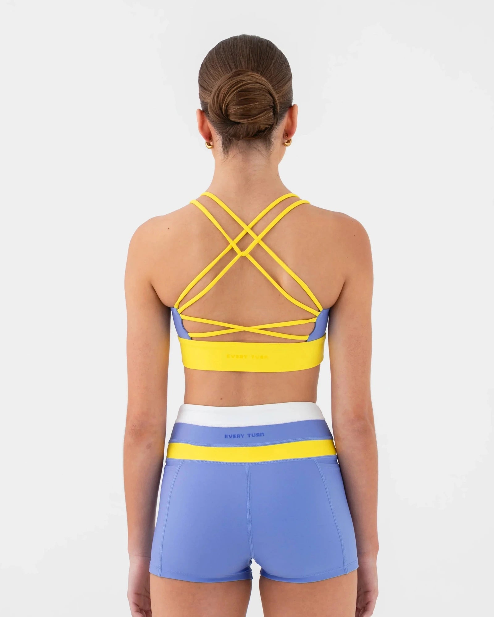 A woman shown from the back wearing a matching dance set consisting of a bra top and shorts. The set features a light blue base color with bright yellow stripes and a white waistband.