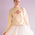 Woman wearing a beige sweatshirt with text and a white skirt on a light purple background