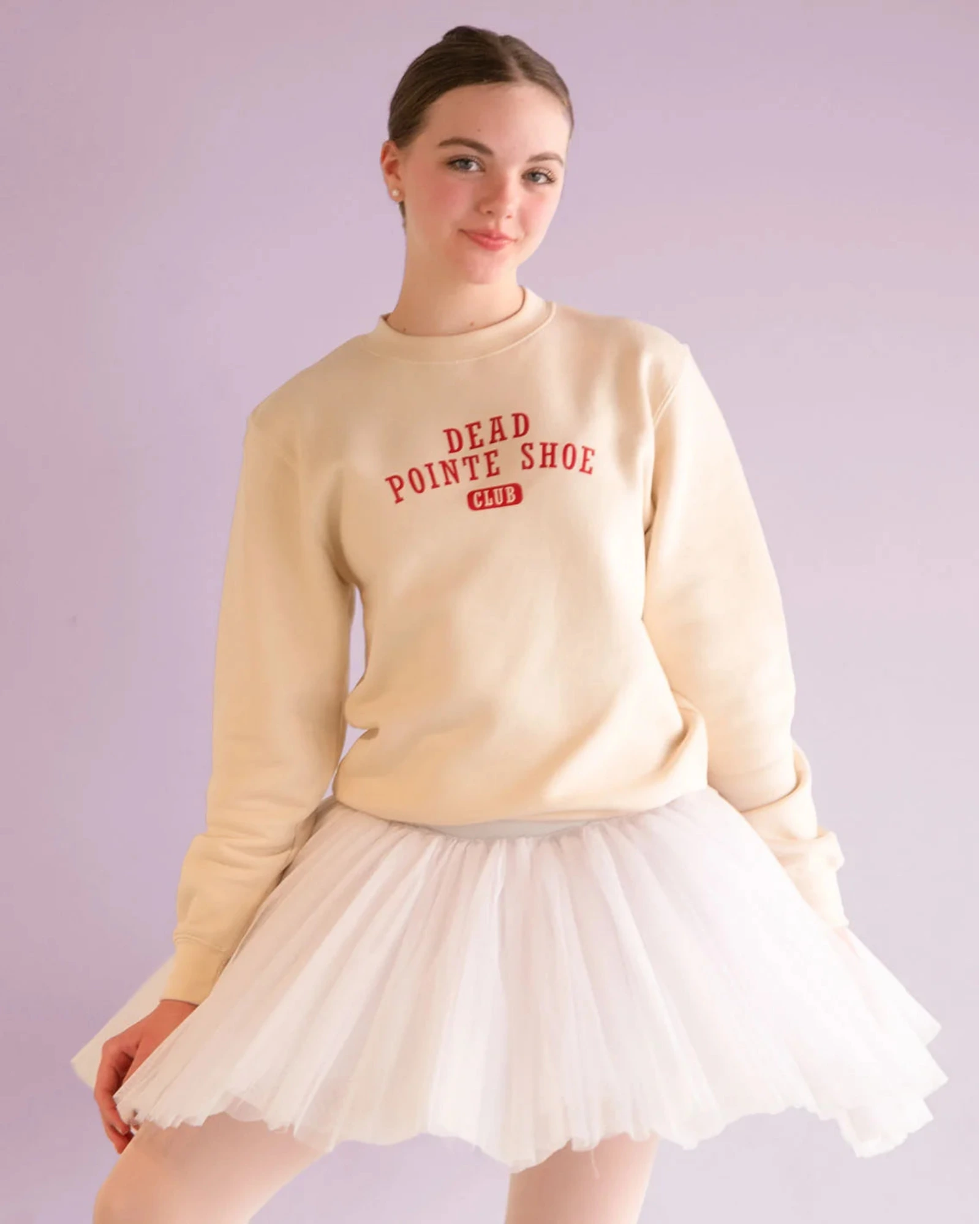 Woman wearing a beige sweatshirt with text and a white skirt on a light purple background