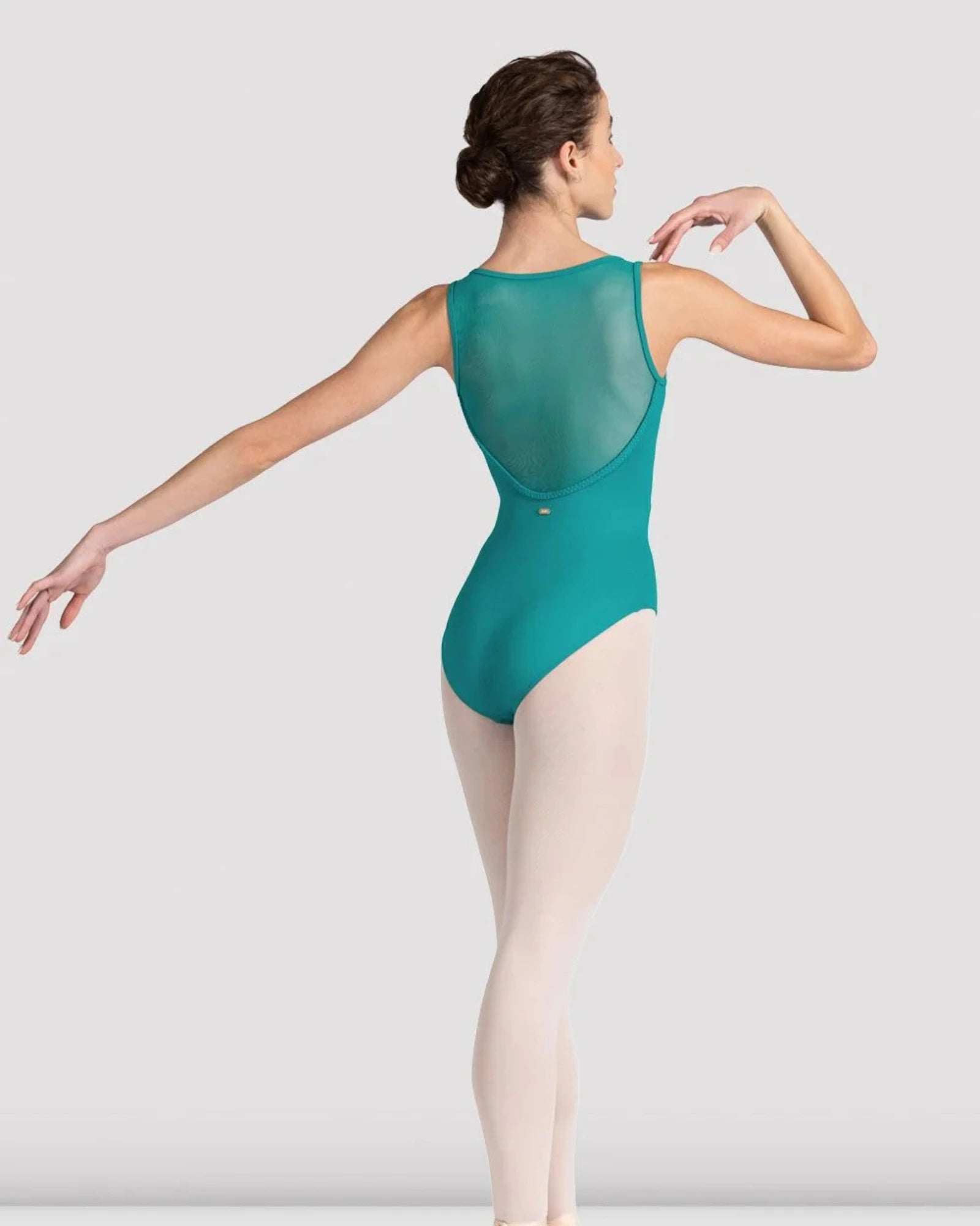 A female dancer shown from the back wearing a teal-green sleeveless leotard with a sheer mesh back panel, paired with light pink tights and pointe shoes, against a light gray studio background.