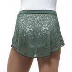 A close-up of a model wearing a sage green lace dance skirt with a matching elastic waistband, paired with a white bodysuit against a plain white background.