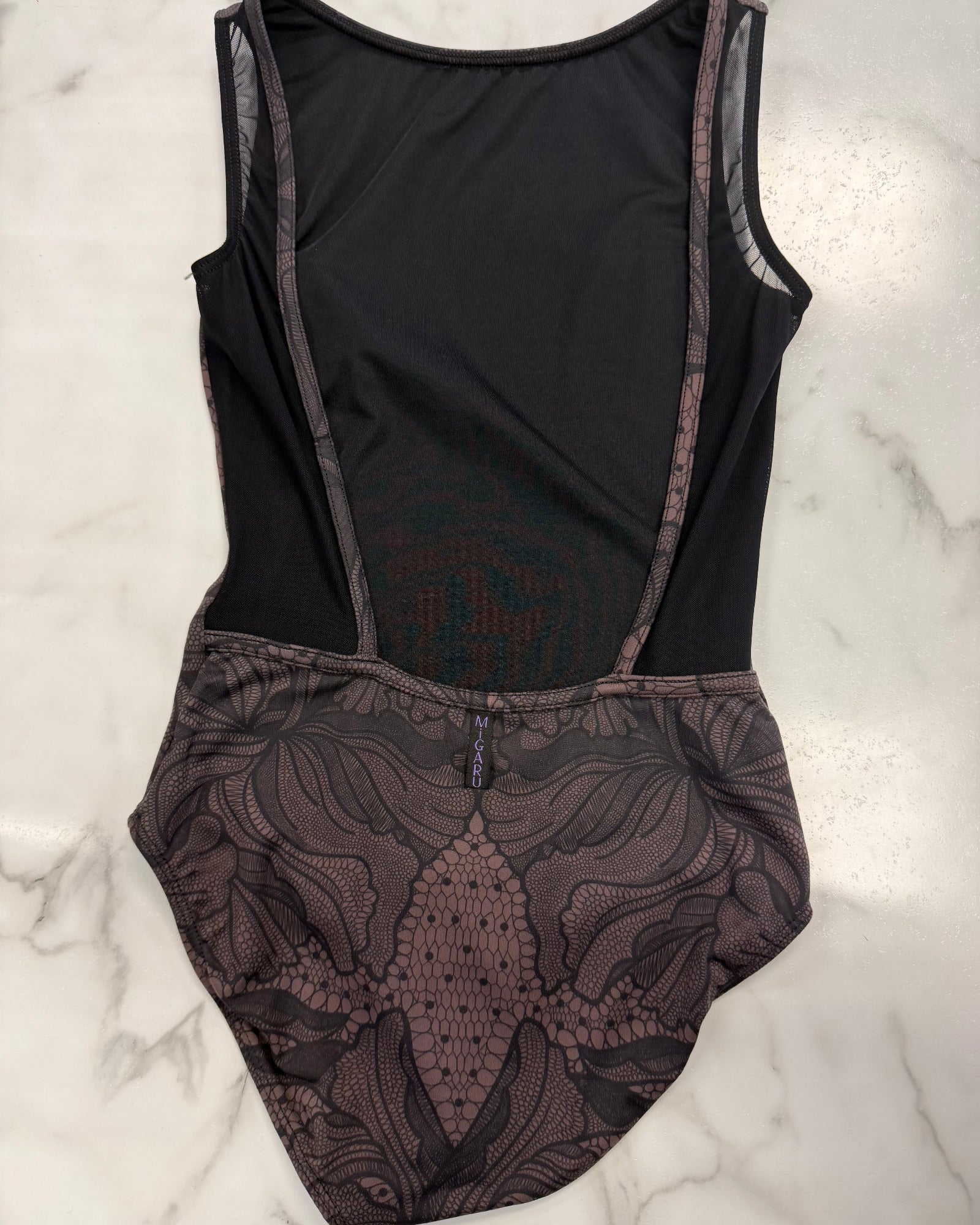 A sleeveless brown leotard with sheer mesh side panels and a brown floral lace overlay on the lower half, laid flat on a white marble surface.