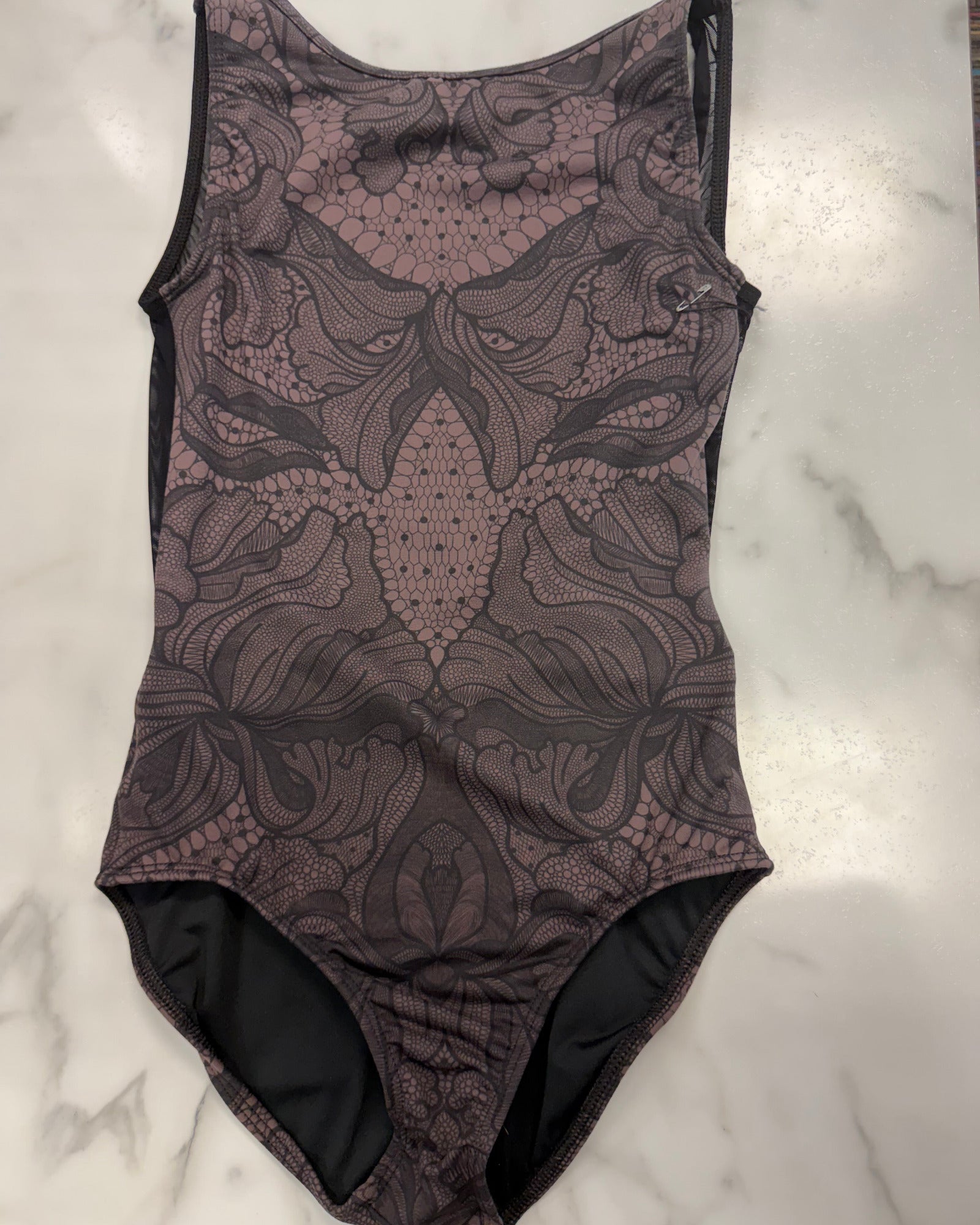 A sleeveless leotard with a dark brown and black abstract floral lace-like pattern, laid flat on a white marble surface.