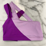A colorblock dance bra in purple and light lavender, laid flat on a white marble surface.