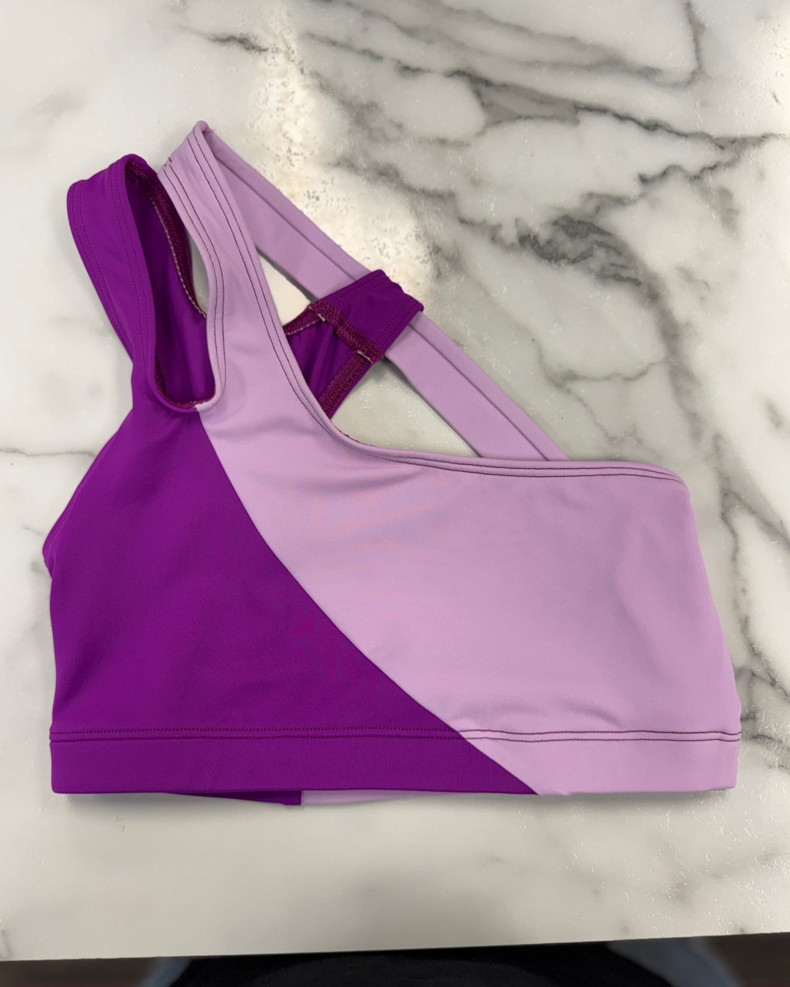 A colorblock dance bra in purple and light lavender, laid flat on a white marble surface.
