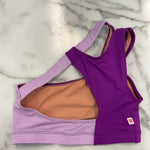 A women's dance bra laid flat on a white marble surface, featuring a split design with a lavender left side, a purple right side, and a peach-colored inner lining.