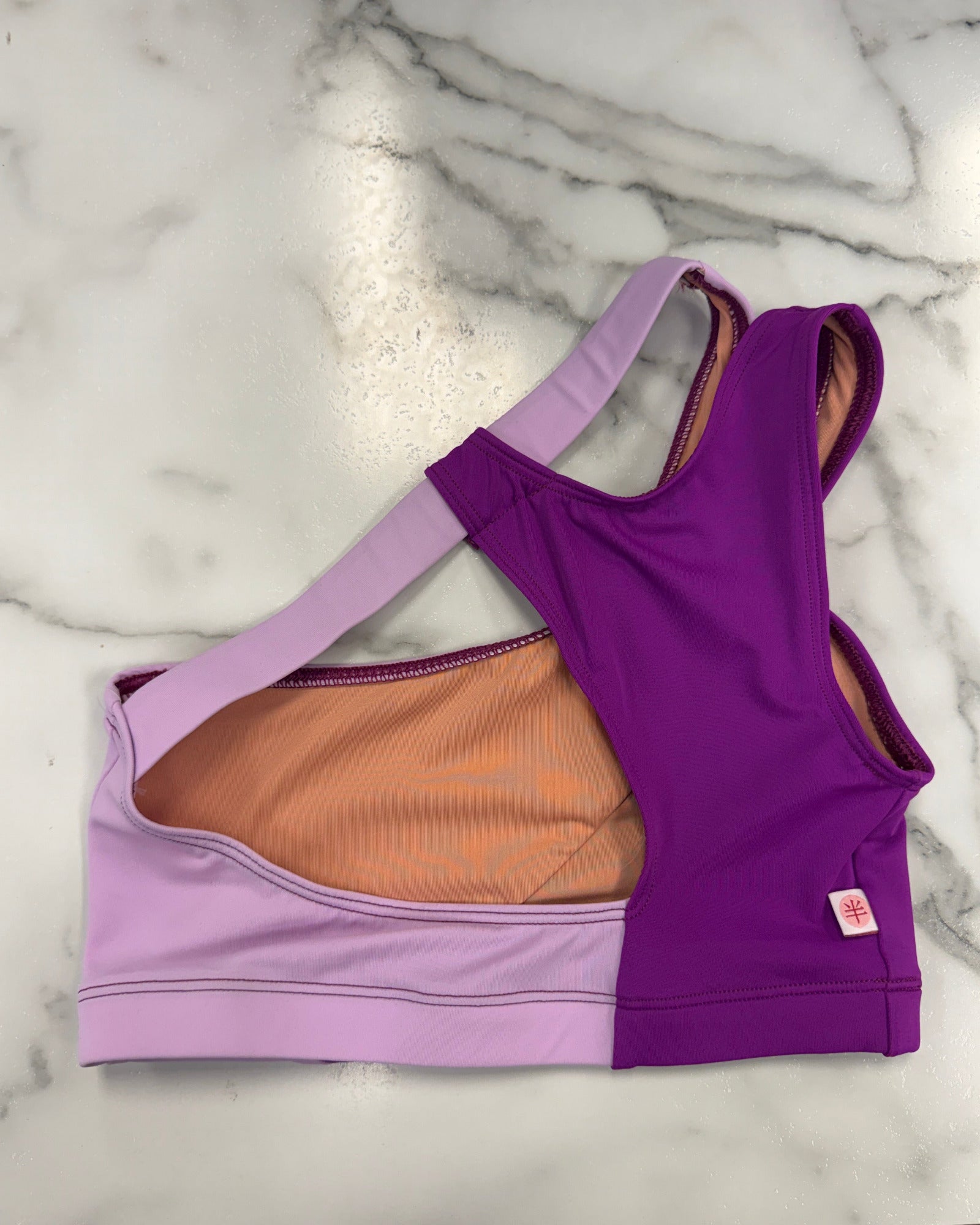A women's dance bra laid flat on a white marble surface, featuring a split design with a lavender left side, a purple right side, and a peach-colored inner lining.