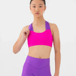 A young female model wearing a bright pink and purple bra top and matching high-waisted shorts against a light gray studio background.