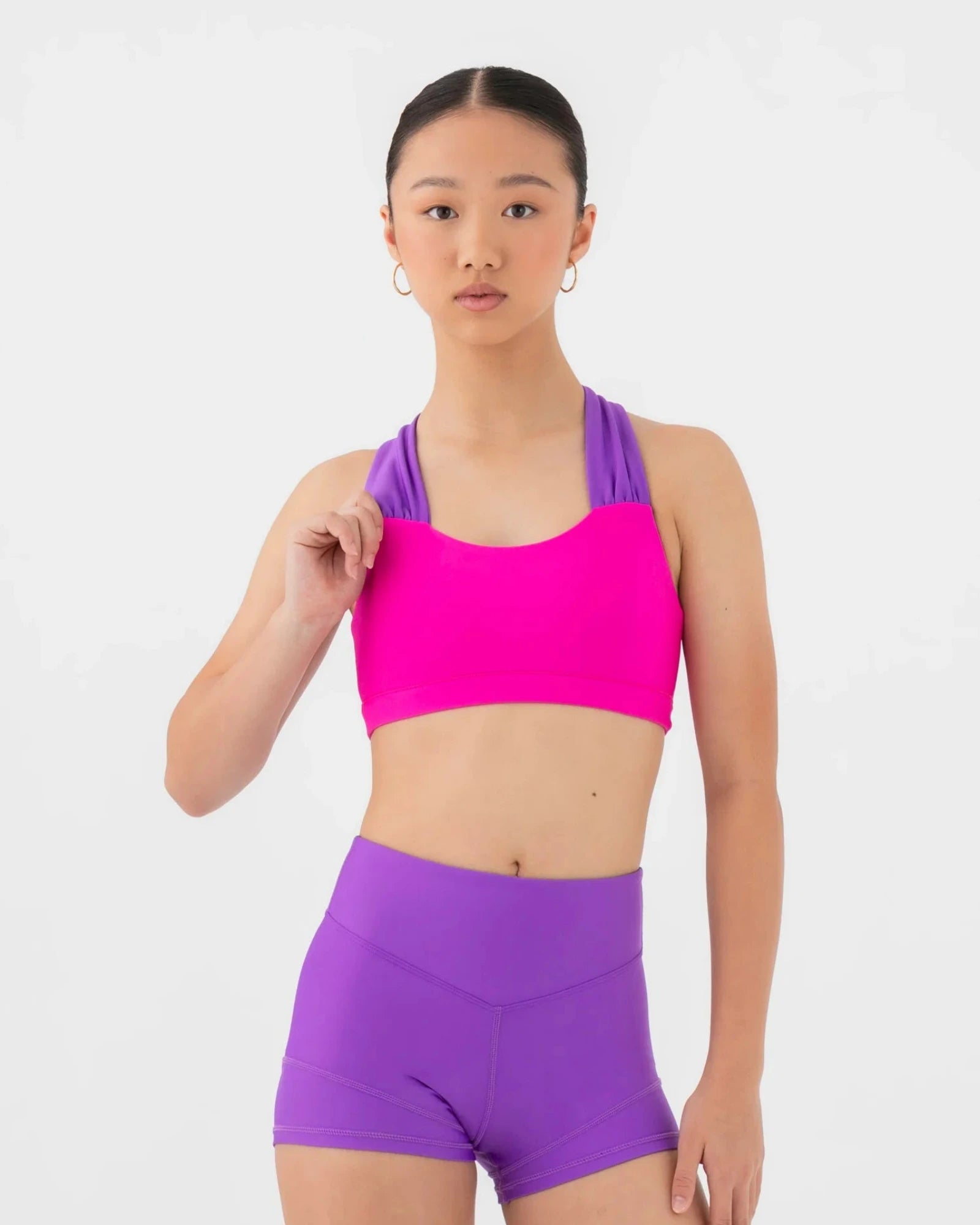 A young female model wearing a bright pink and purple bra top and matching high-waisted shorts against a light gray studio background.