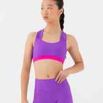 A young woman modeling a matching purple dance set consisting of a bra top with a bright pink band and high-waisted shorts, set against a plain light gray background.