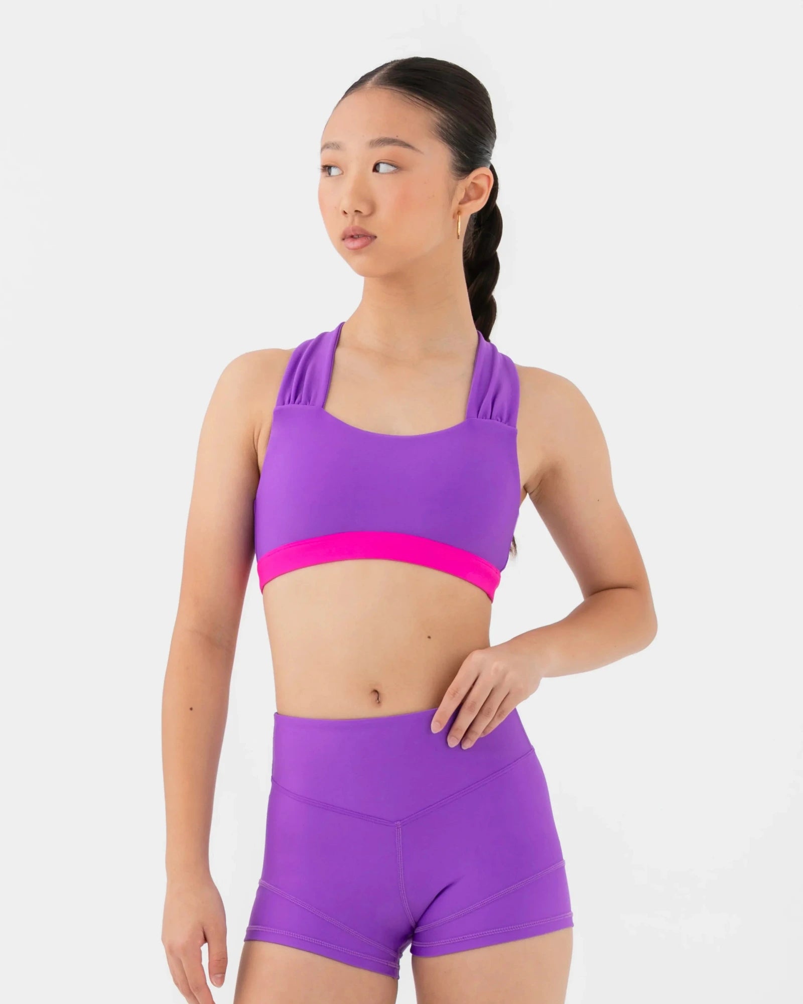 A young woman modeling a matching purple dance set consisting of a bra top with a bright pink band and high-waisted shorts, set against a plain light gray background.