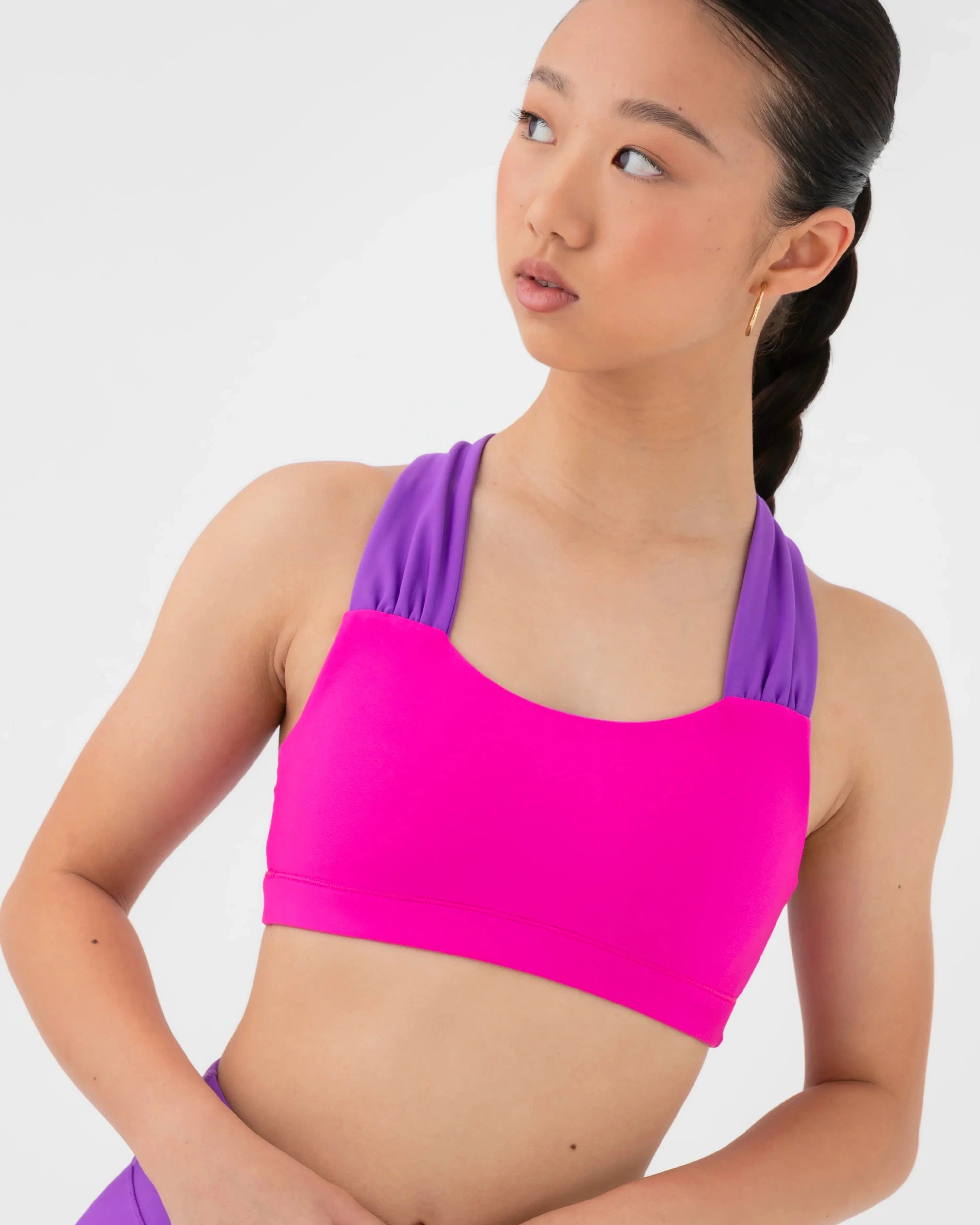 A woman modeling a bright pink and purple bra top and matching purple shorts against a light gray studio background.