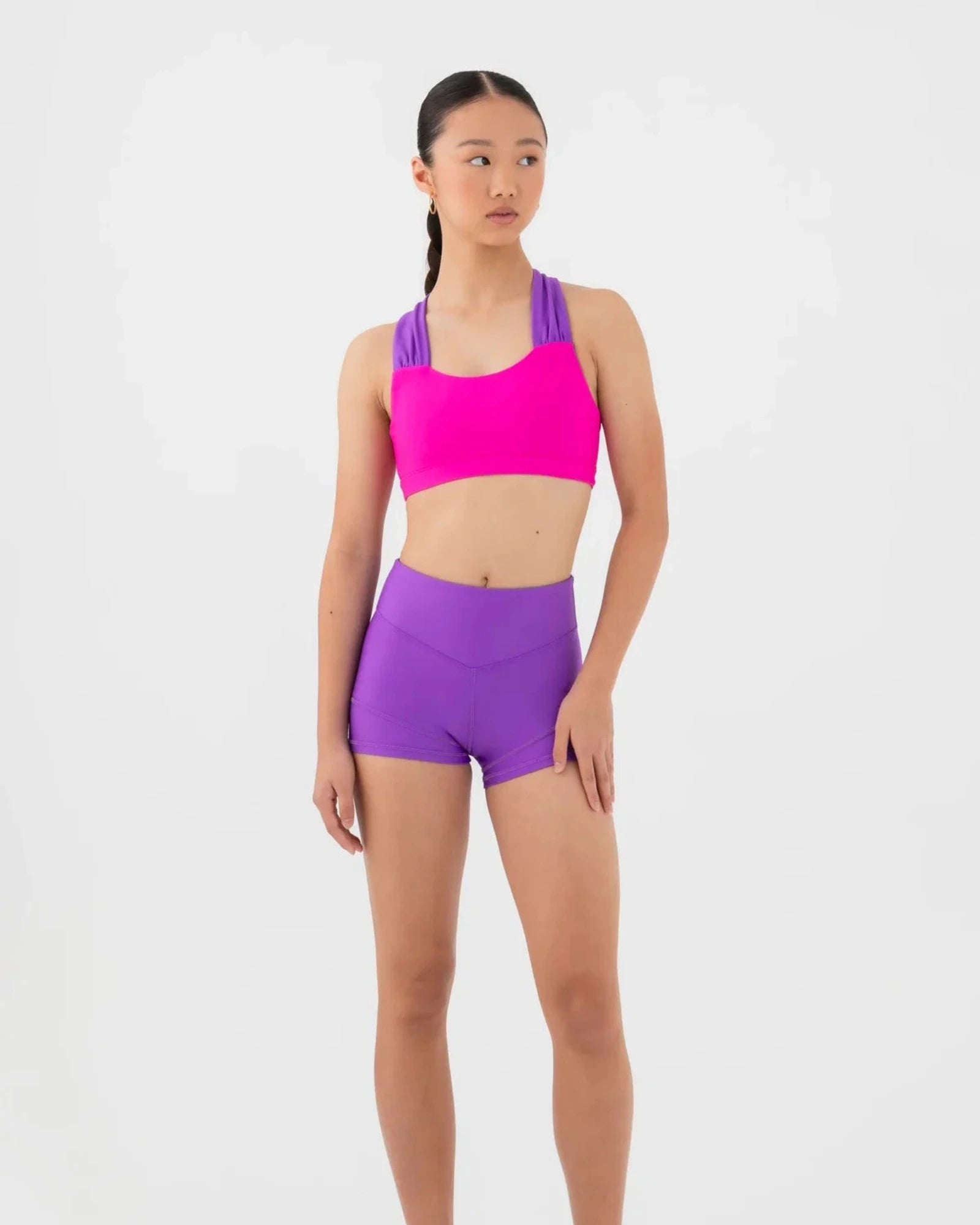 A woman modeling a bright pink bra top and purple dance shorts against a plain white studio background.