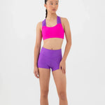 A woman modeling a bright pink bra top and purple dance shorts against a plain white studio background.