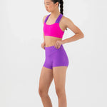A woman modeling a bright pink bra top and purple high-waisted dance shorts against a light gray studio background.