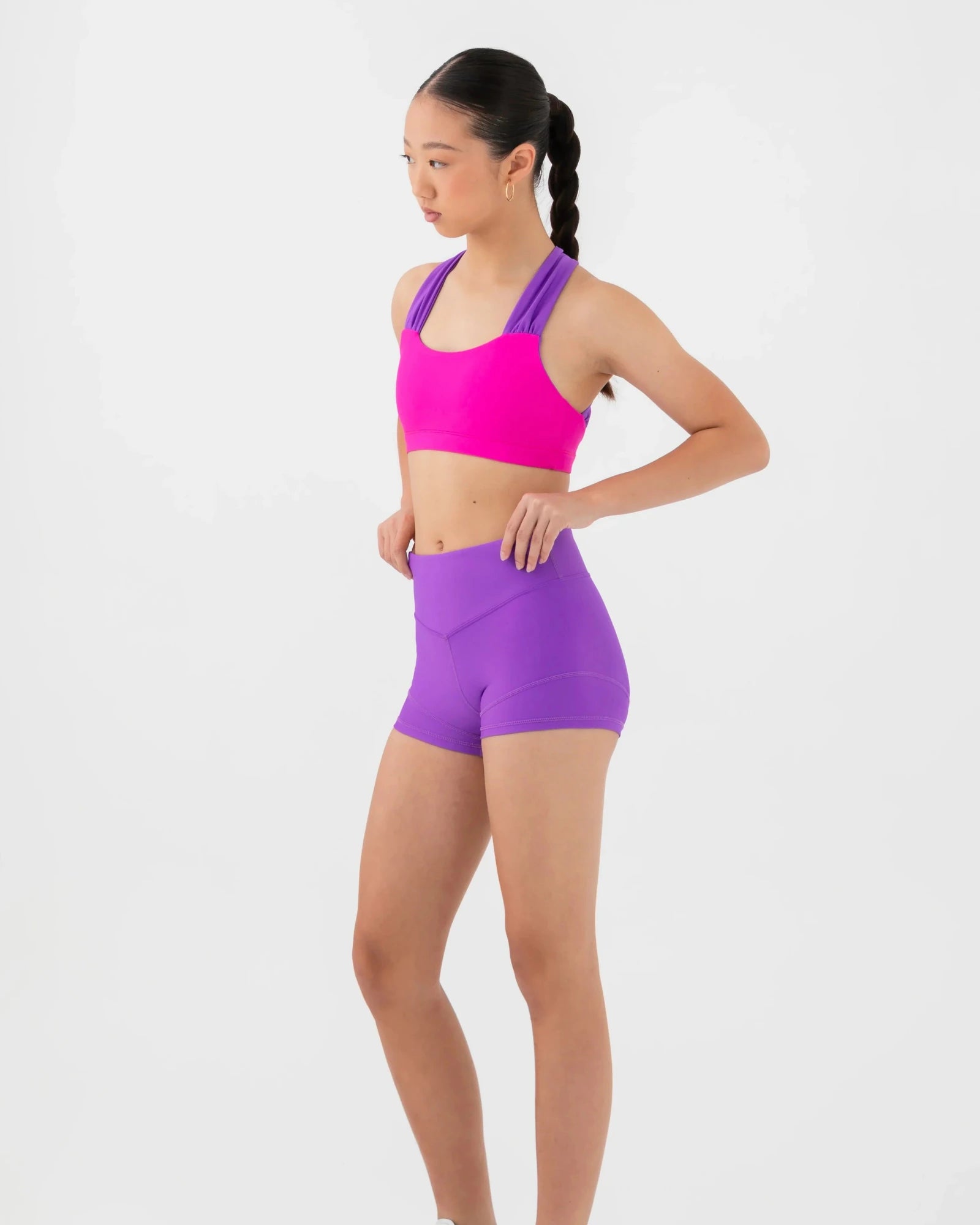A woman modeling a bright pink bra top and purple high-waisted dance shorts against a light gray studio background.