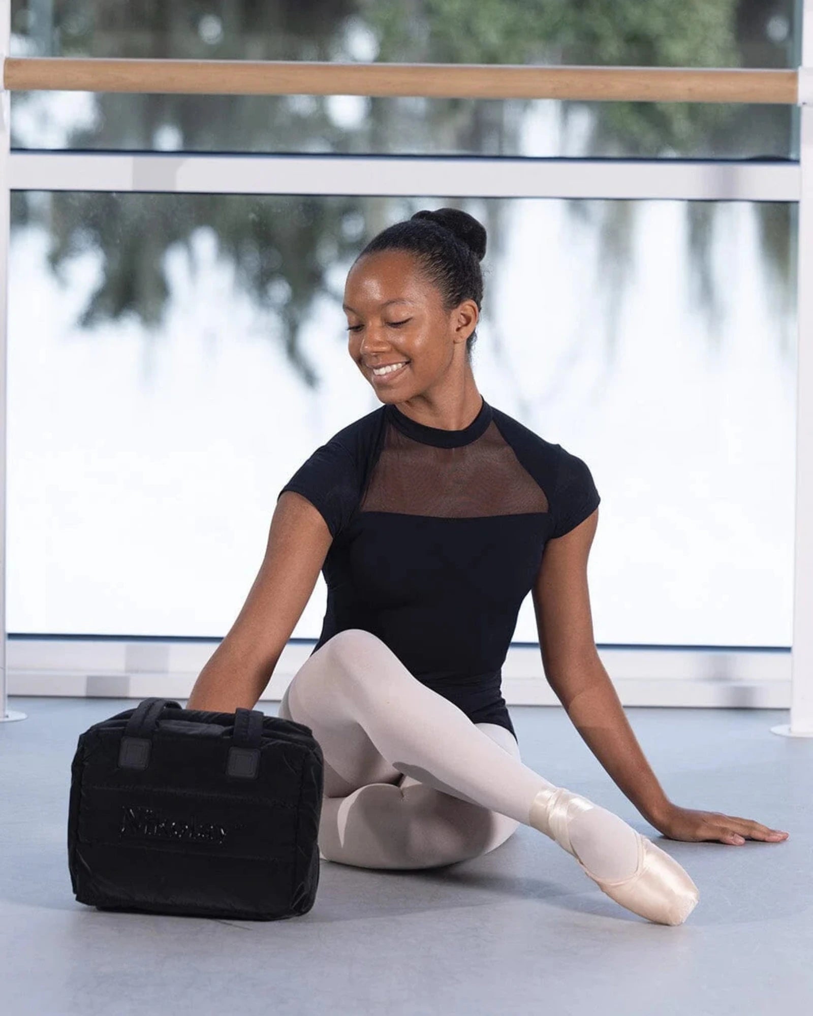 Ballerina sitting on the floor with a black bag in a ballet studio.
