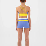A woman viewed from the back wearing a matching dance set consisting of a yellow and blue strappy bra top and blue shorts with a yellow and white waistband on a light gray studio background.