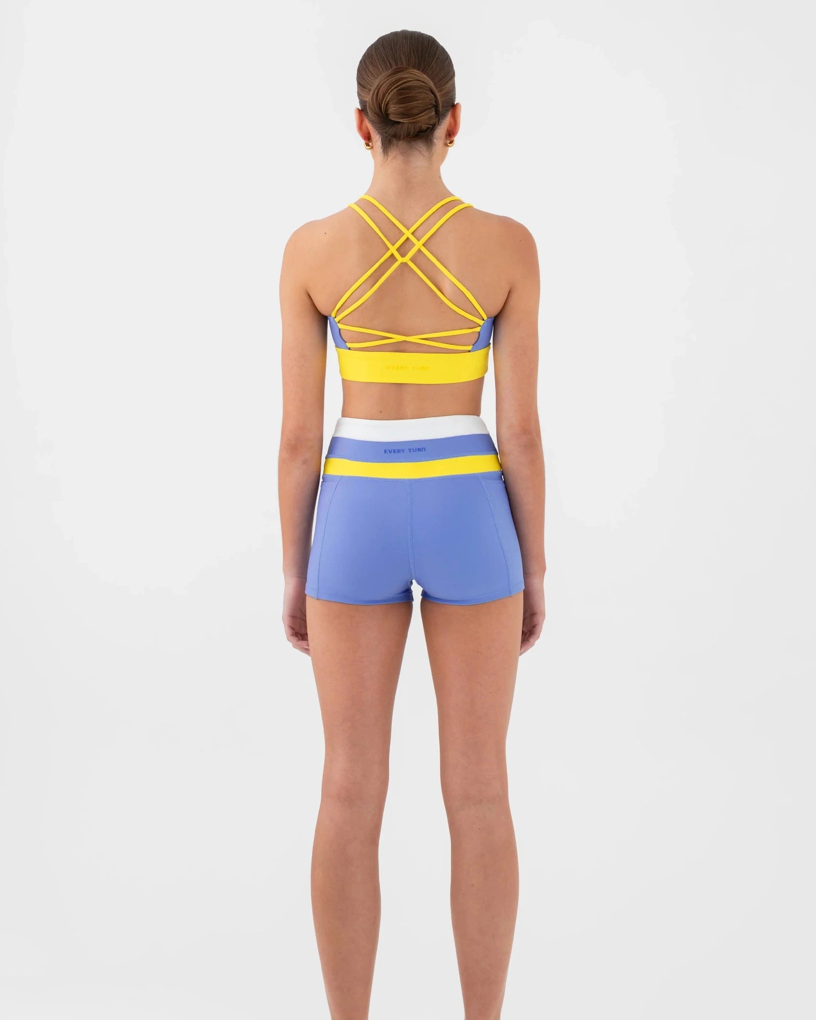 A woman viewed from the back wearing a matching dance set consisting of a yellow and blue strappy bra top and blue shorts with a yellow and white waistband on a light gray studio background.