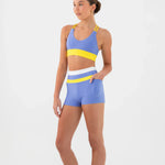 A woman modeling a matching athletic set consisting of a blue bra top with yellow and white stripes, and matching blue shorts with a white waistband, on a plain white background.