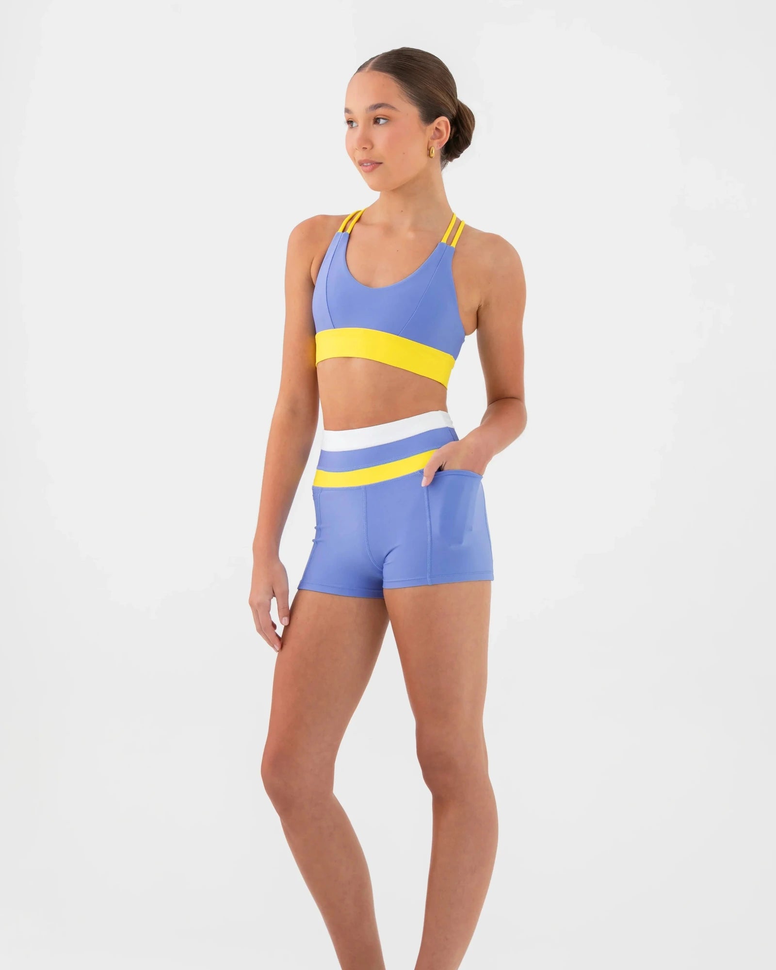 A woman modeling a matching athletic set consisting of a blue bra top with yellow and white stripes, and matching blue shorts with a white waistband, on a plain white background.