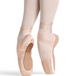 A close-up of a dancer's legs in pale pink tights, wearing light pink satin pointe shoes, posed en pointe against a white background.