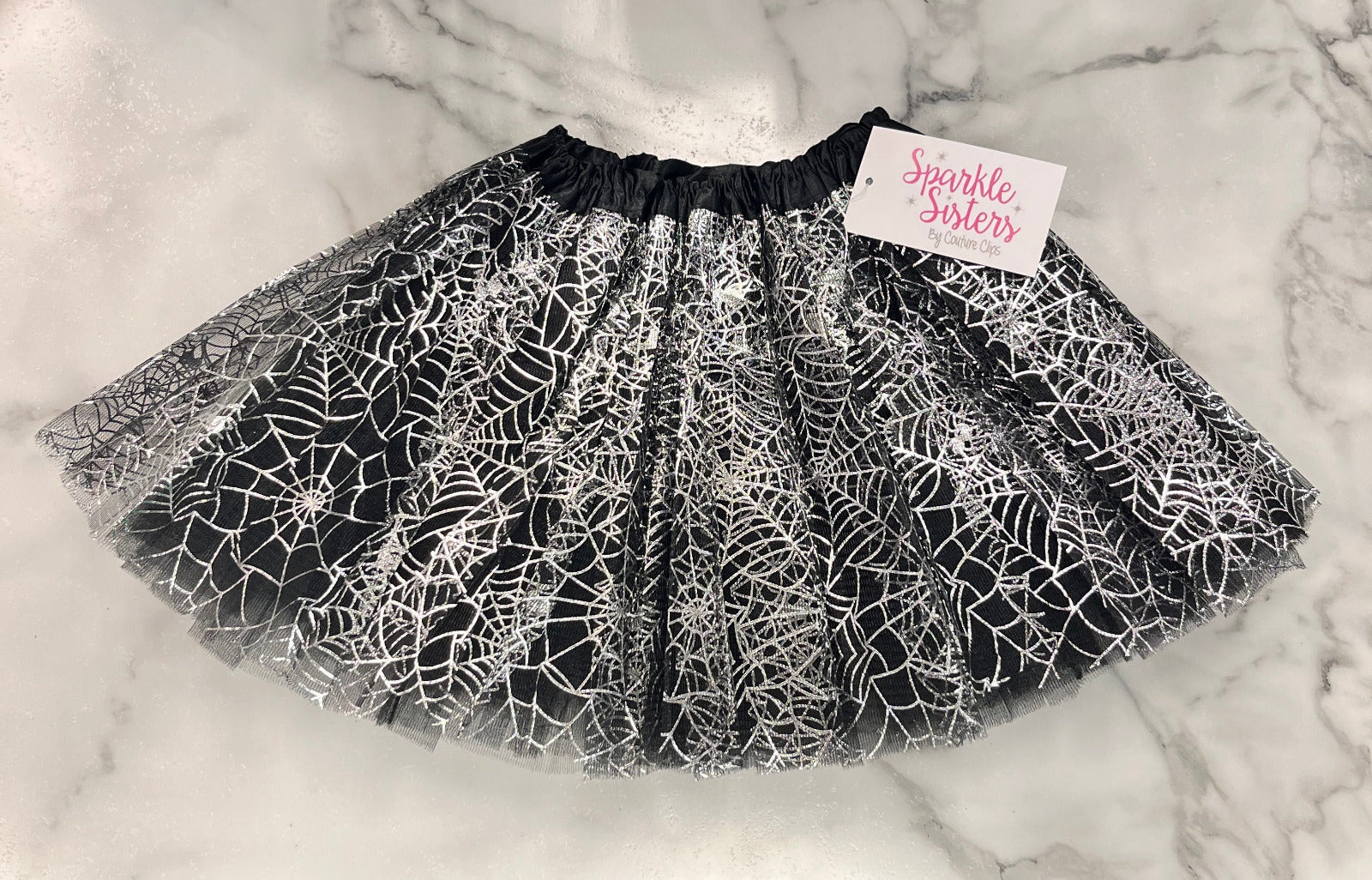 A black and white tulle tutu skirt with a spiderweb pattern, featuring a black elastic waistband and a white paper tag that reads 'Sparkle Sisters' in pink script, laid flat on a white marble surface.