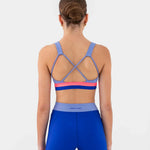 A woman viewed from the back wearing a blue dance bra with purple and pink accents and matching blue athletic shorts against a light gray studio background.