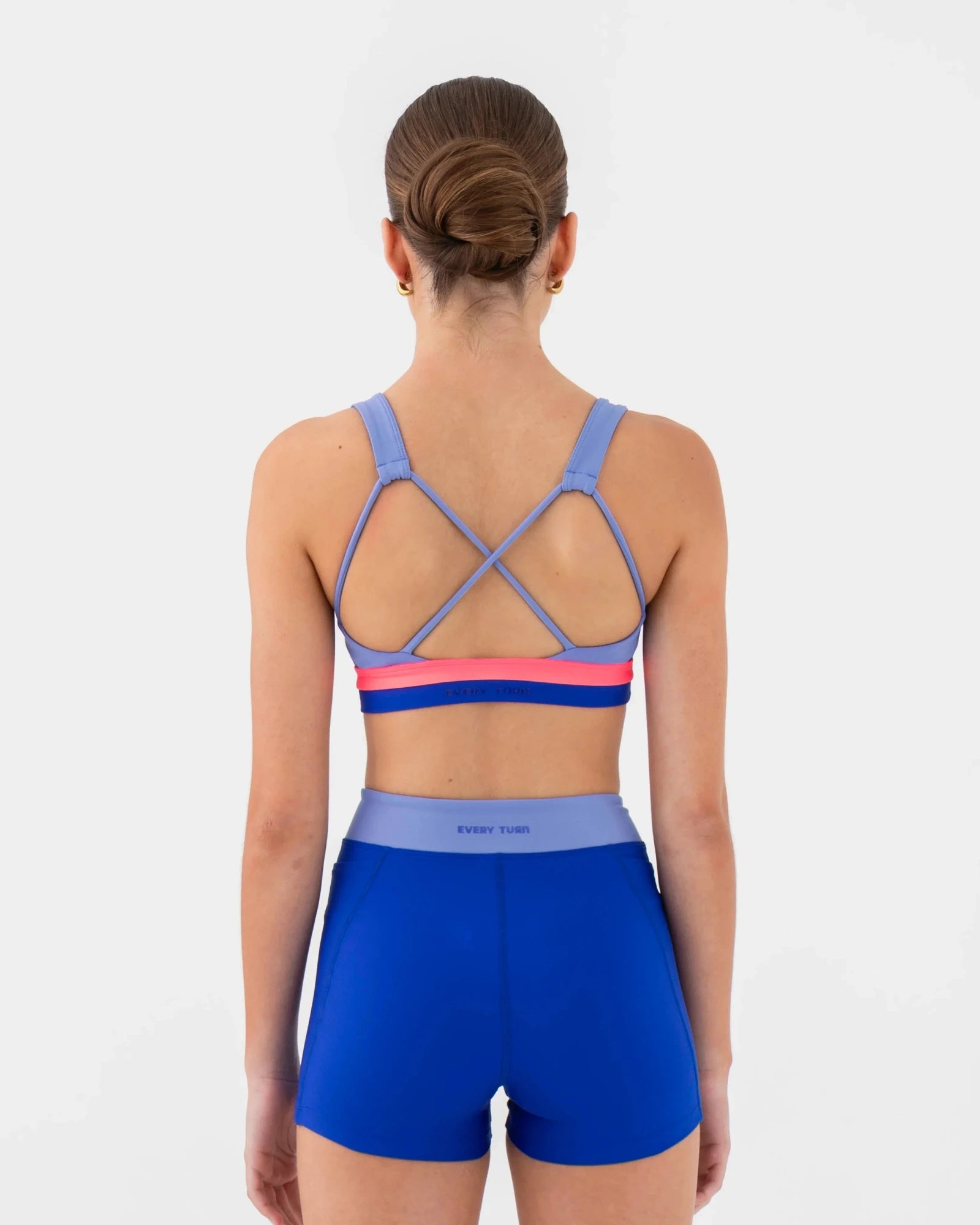 A woman viewed from the back wearing a blue dance bra with purple and pink accents and matching blue athletic shorts against a light gray studio background.