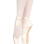 A pair of satin pointe shoes worn on a dancer's feet, shown in a side profile against a solid white background. The shoes are light pink with white satin ribbons and white tights.