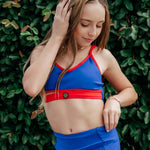 A young woman wearing a blue and red dance bra and matching blue shorts, posing in front of a dense green leaf wall.