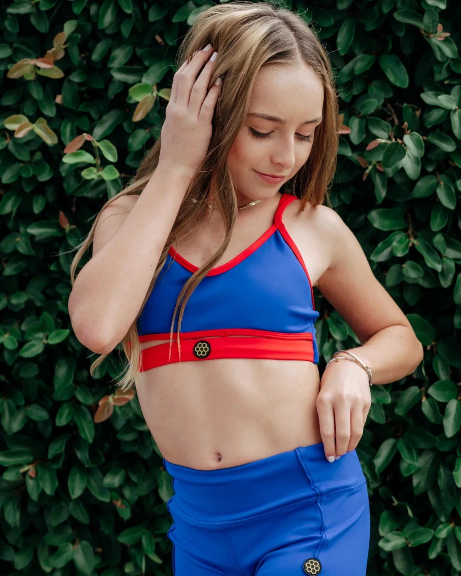 A young woman wearing a blue and red dance bra and matching blue shorts, posing in front of a dense green leaf wall.