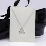 A silver-tone necklace with a tap shoe tap pendant displayed on a white card, set against a background of black ballet flats with black ribbons.