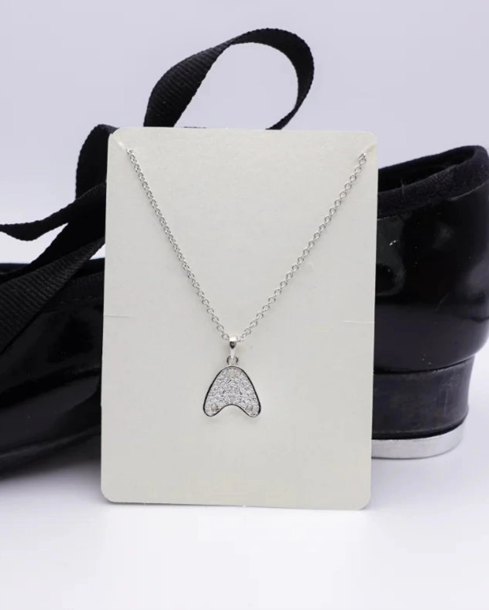 A silver-tone necklace with a tap shoe tap pendant displayed on a white card, set against a background of black ballet flats with black ribbons.