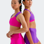 Two young female athletes posing back-to-back in matching dance wear. The girl on the left wears a bright magenta bra top and shorts, while the girl on the right wears a purple bra top and matching purple high-waisted shorts.