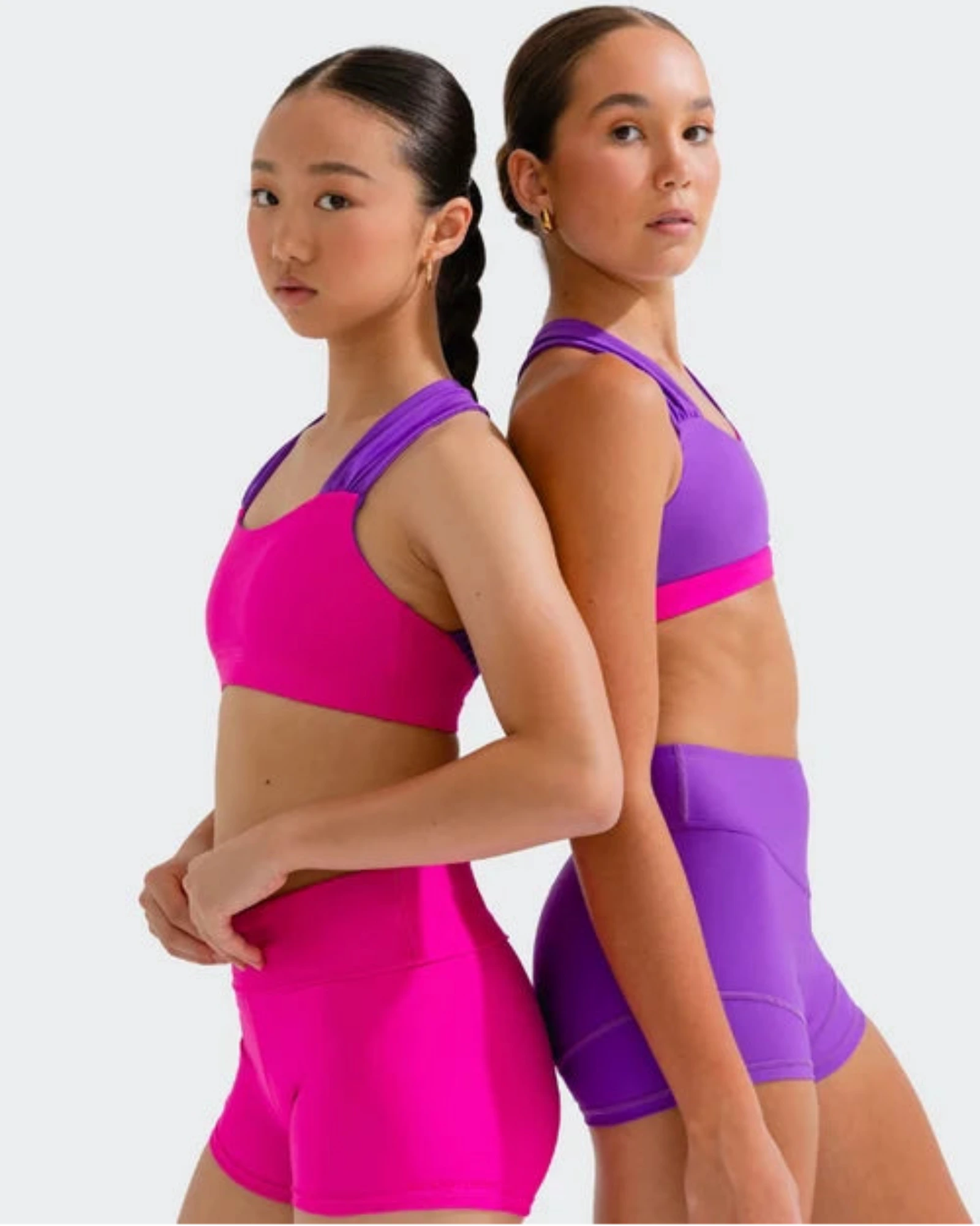 Two young female athletes posing back-to-back in matching dance wear. The girl on the left wears a bright magenta bra top and shorts, while the girl on the right wears a purple bra top and matching purple high-waisted shorts.