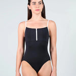 A woman modeling a black leotard with thin white shoulder straps and a white vertical zipper detail at the center of the chest, set against a plain light gray studio background.