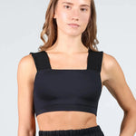 A woman modeling a black dance bra with wide, padded shoulder straps and a square neckline, paired with black high-waisted shorts, against a light gray studio background.