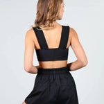A woman shown from the back wearing a black dance crop top with wide shoulder straps and matching black high-waisted shorts against a light gray studio background.