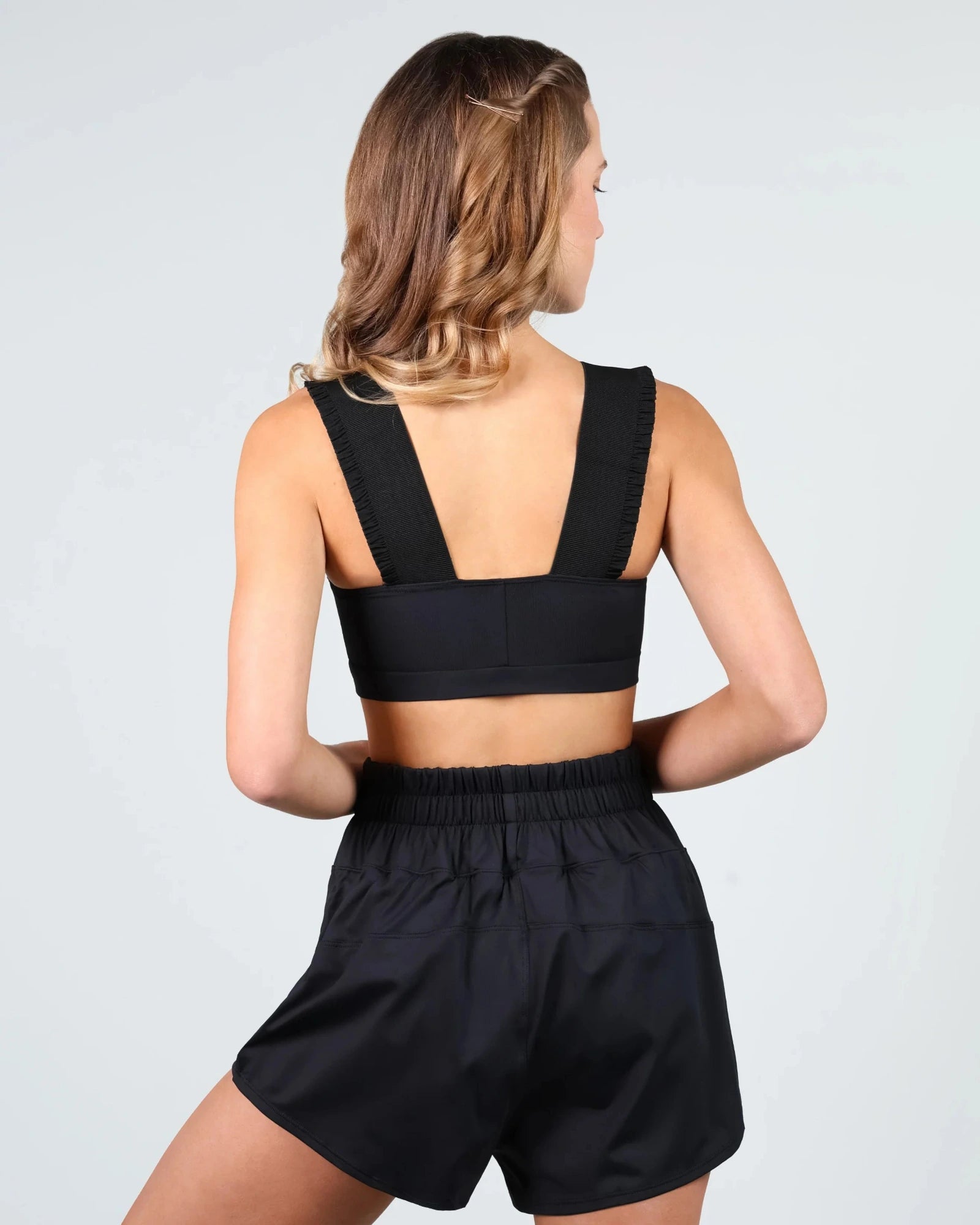 A woman shown from the back wearing a black dance crop top with wide shoulder straps and matching black high-waisted shorts against a light gray studio background.
