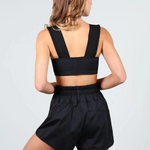 A woman shown from the back wearing a dance crop top with wide shoulder straps and matching black high-waisted shorts against a light gray studio background.