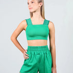 A young woman modeling a matching green dance set consisting of a ribbed crop top with square neckline and wide shoulder straps, paired with high-waisted elastic-waist shorts, against a light gray studio background