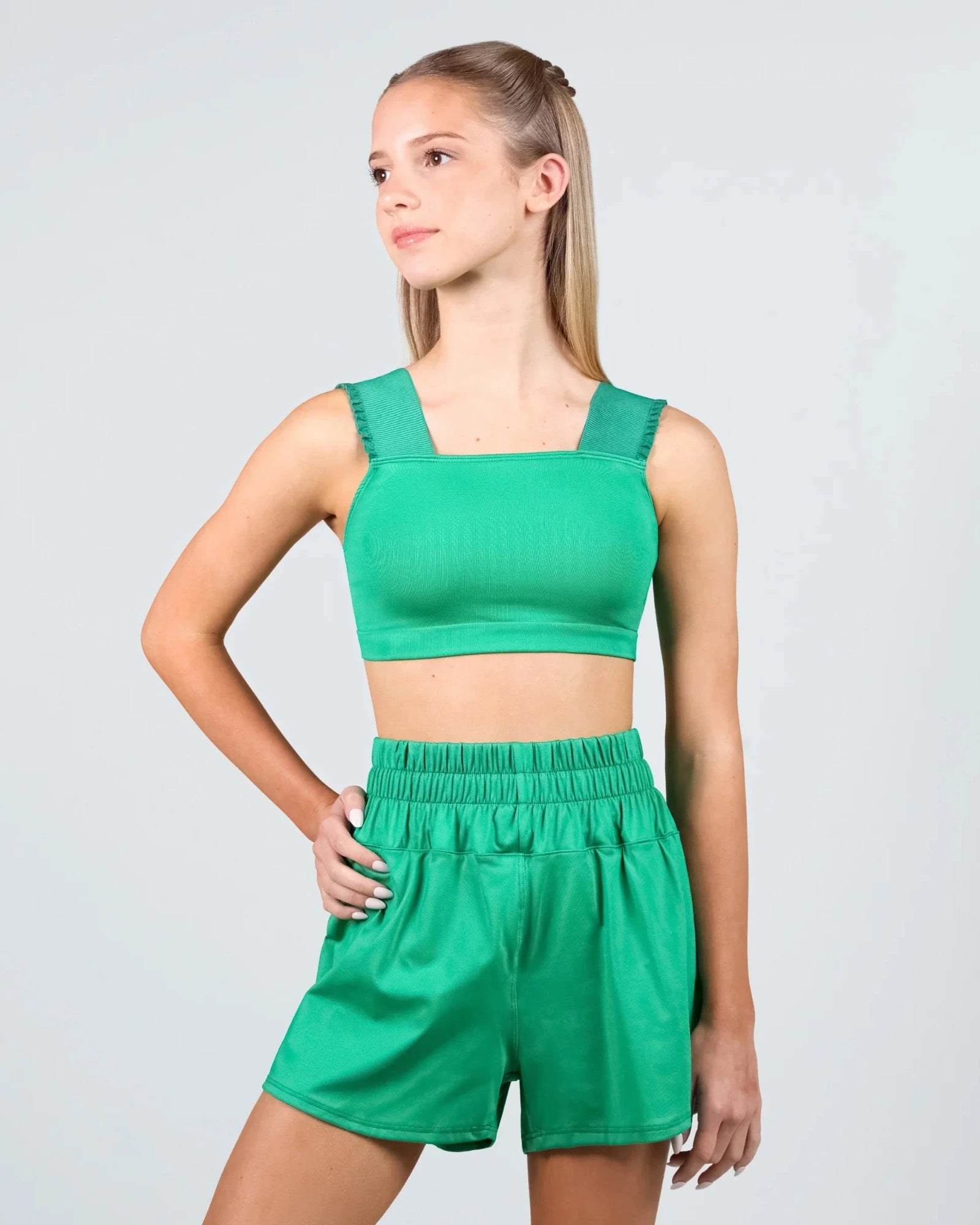 A young woman modeling a matching green dance set consisting of a ribbed crop top with square neckline and wide shoulder straps, paired with high-waisted elastic-waist shorts, against a light gray studio background