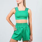 A young woman modeling a matching green dance set consisting of a ribbed crop top with square neckline and wide shoulder straps, paired with high-waisted elastic-waist shorts, against a light gray studio background.