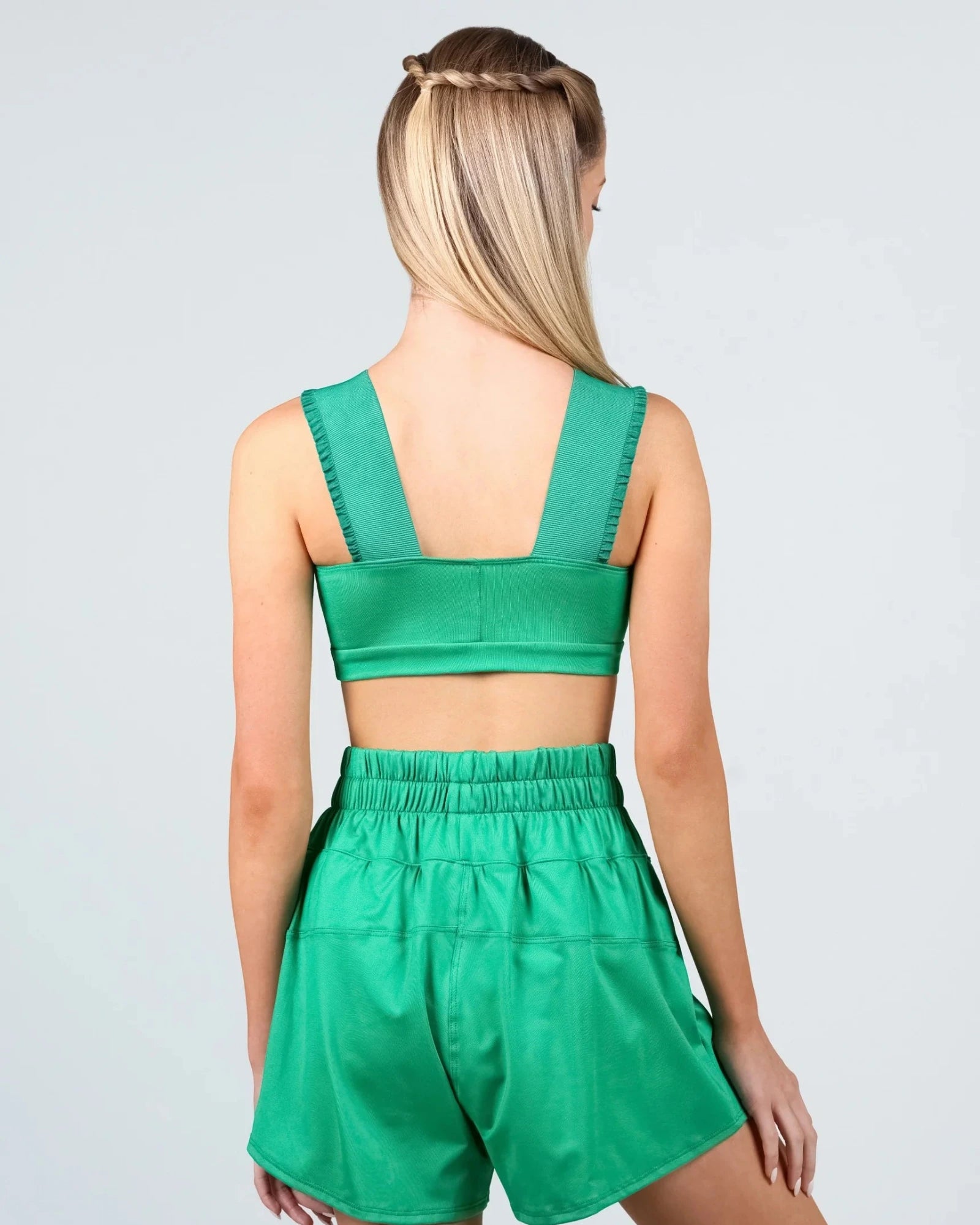 A woman shown from the back wearing a matching green two-piece dance outfit consisting of a crop top and high-waisted shorts, against a light gray studio background.