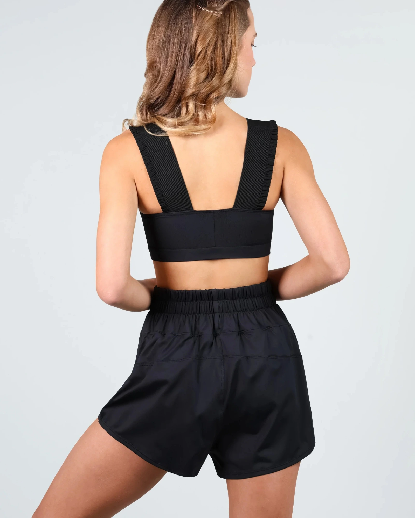 A woman shown from the back wearing a black dance crop top with wide shoulder straps and matching black high-waisted shorts against a light gray studio background.