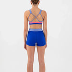 A woman viewed from the back wearing a matching dance set: a bra top with crisscross straps and a blue waistband, plus fitted blue shorts. The  bra top has thin pink and red horizontal bands near the back.