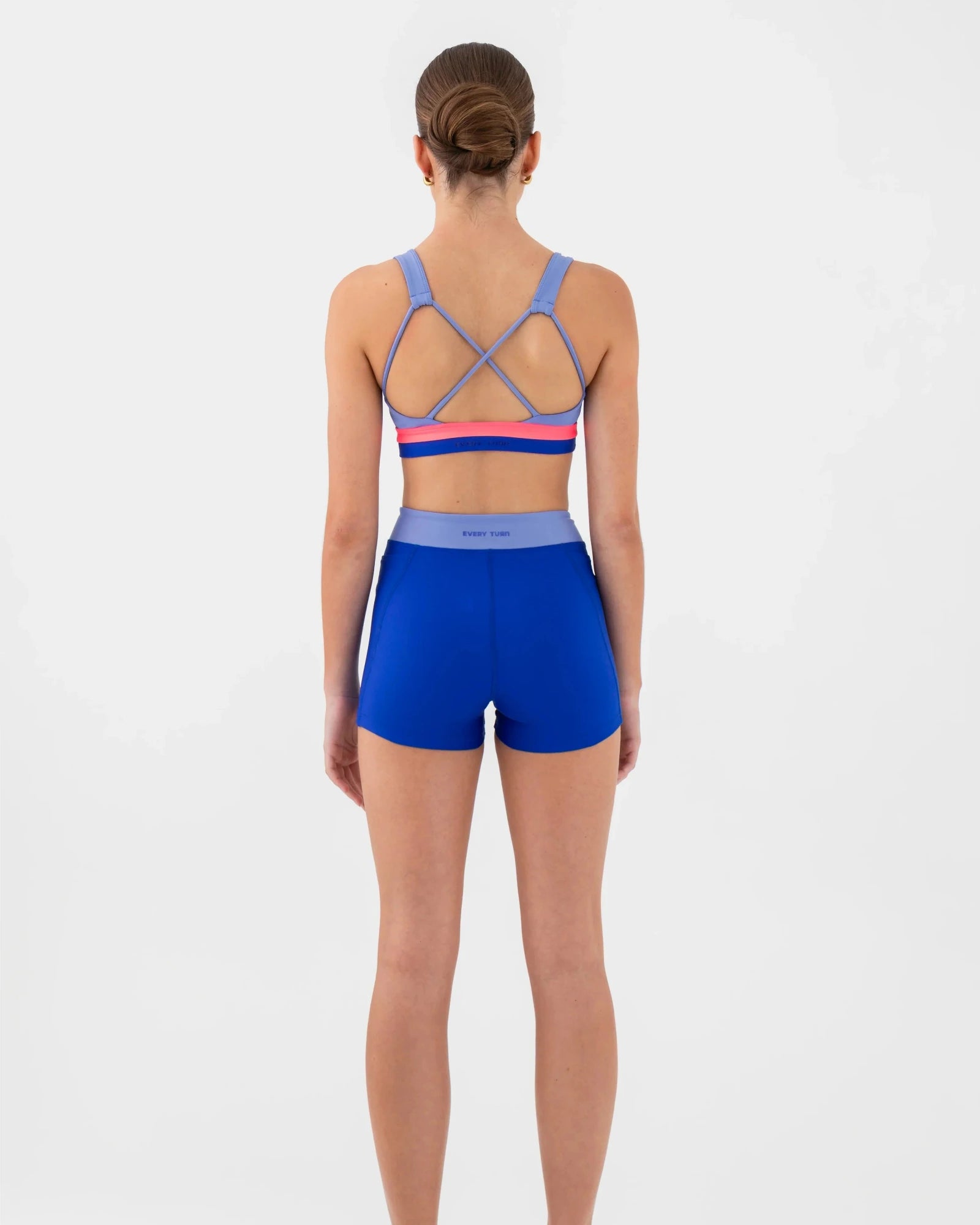A woman viewed from the back wearing a matching dance set: a bra top with crisscross straps and a blue waistband, plus fitted blue shorts. The  bra top has thin pink and red horizontal bands near the back.